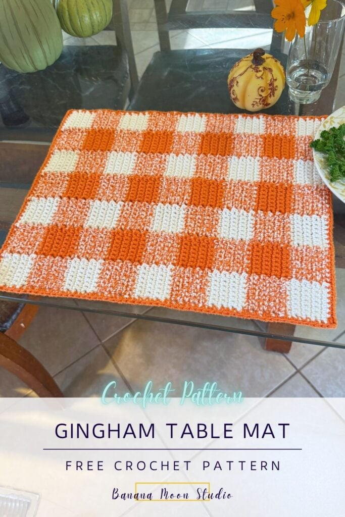 Crochet gingham placemat pattern lying on a glass table top with other decor items. A panel with text reads: Gingham table mat free crochet pattern, banana moon studio.