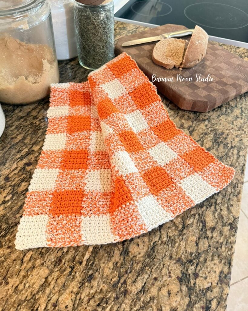 Gingham fall crochet kitchen dowel lying folded on a granite countertop along with a loaf of bread on a cutting board. Text on the image reads: Banana moon studio.