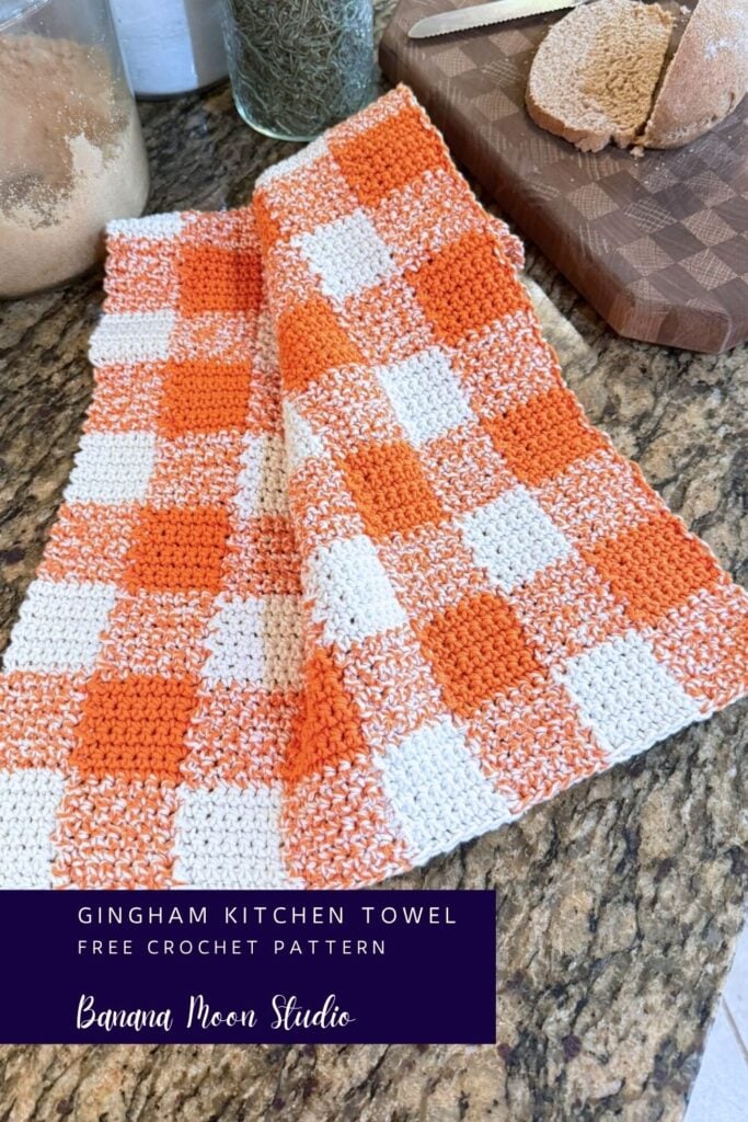 A folded crochet gingham kitchen towel lying on a granite countertop next to a loaf of bread on a cutting board. A panel with text reads: Gingham kitchen towel free crochet pattern, banana moon studio.