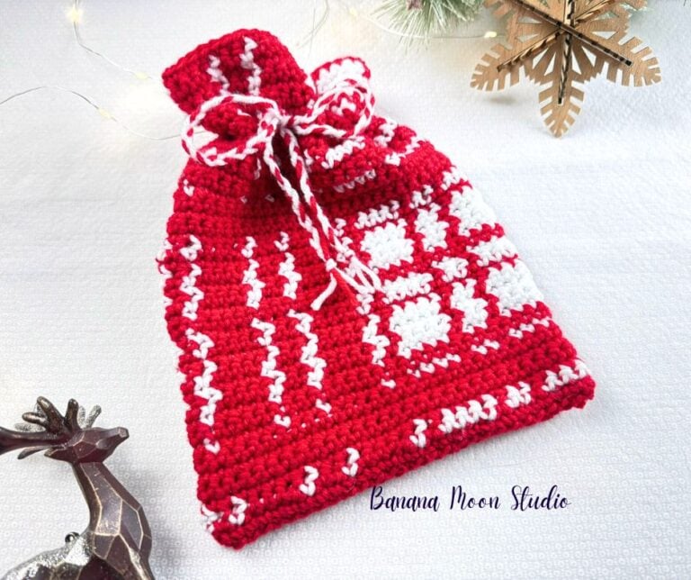 Red and white plaid Christmas crochet drawstring bag lying on a white surface with Christmas decorations. Text on the image reads: Banana Moon Studio.