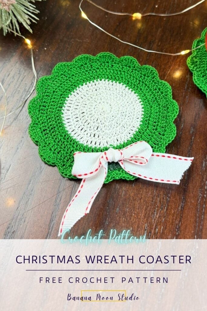 Crochet Christmas wreath shaped drink coaster lying on a wooden surface along with string lights. A panel with text reads: Christmas wreath coaster free crochet pattern, banana moon studio.