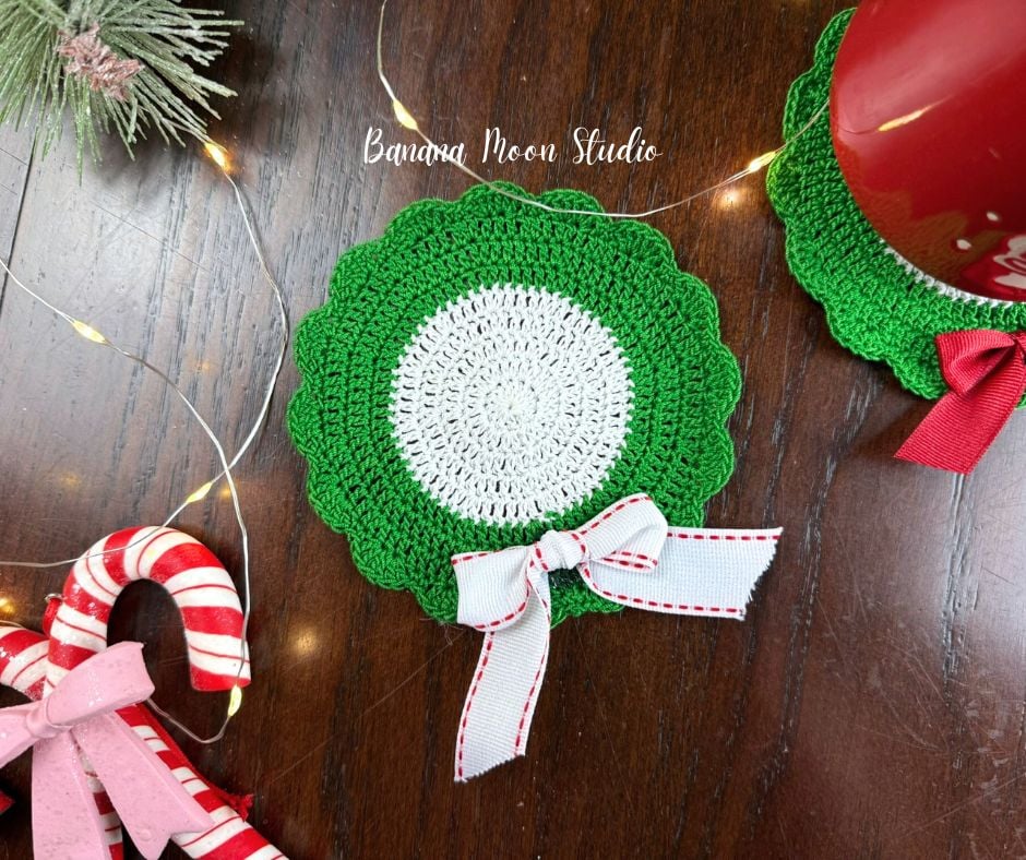 Christmas crochet coaster to look like a wreath lying flat on a wooden surface along with other Christmas decor. Text on the image reads: Banana Moon Studio.