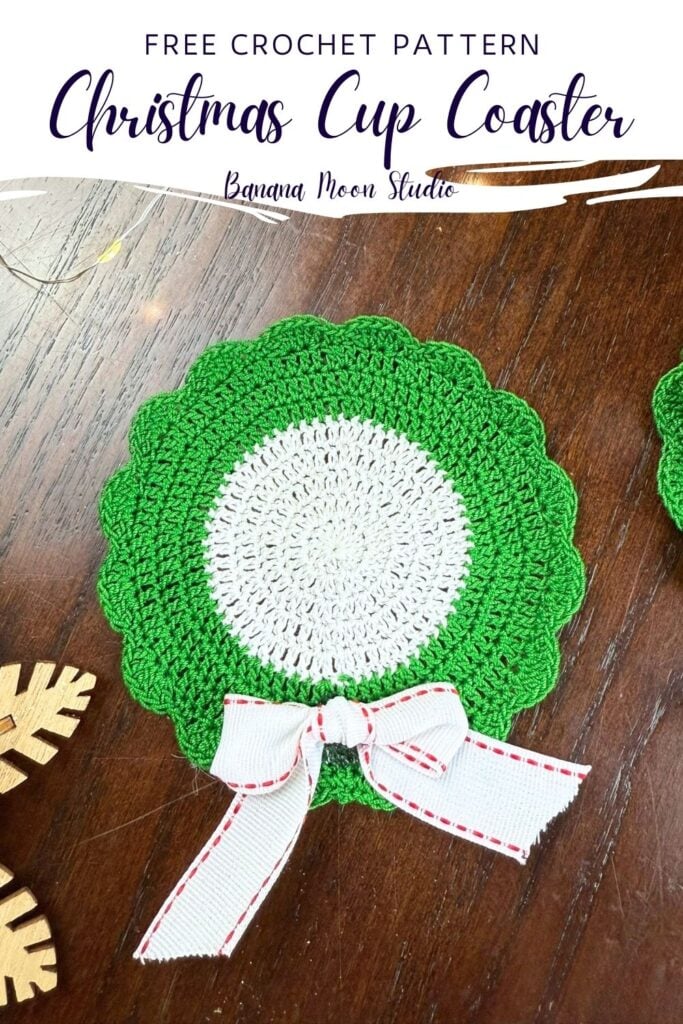 Christmas crochet coaster that looks like a wreath lying flat on a wooden surface. A panel with text reads: Free crochet pattern, christmas cup coaster, banana moon studio.