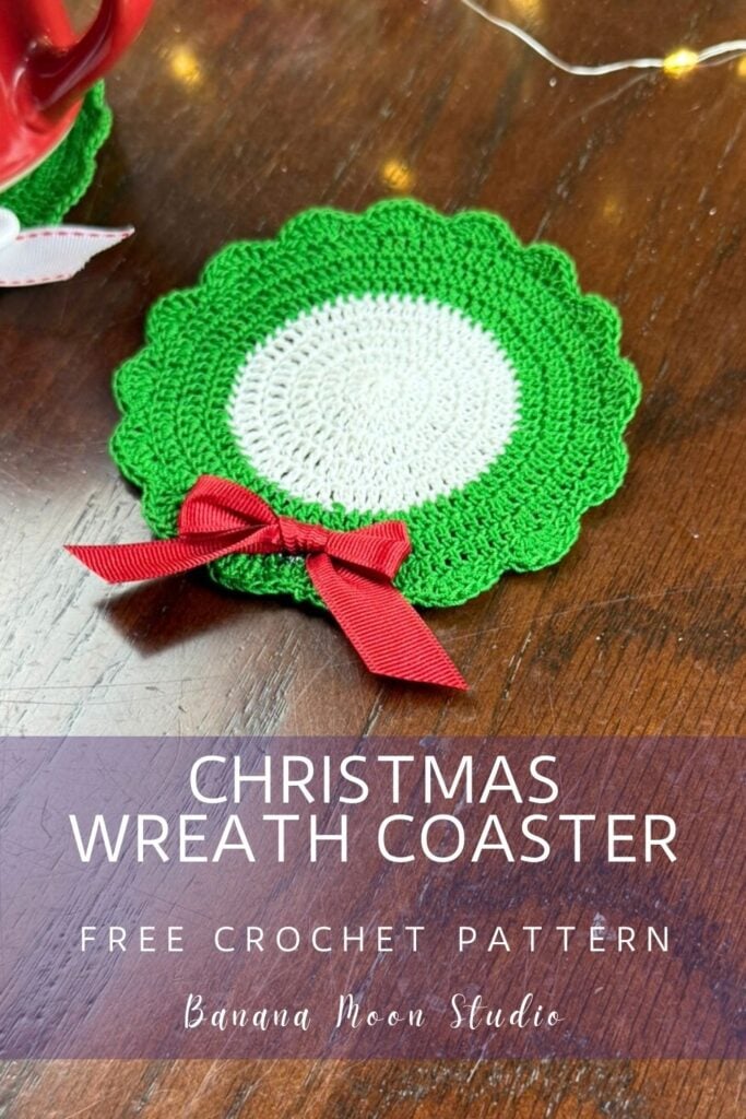 Crochet Christmas coaster that looks like a wreath lying flat on a wooden surface. A panel with text reads: Christmas wreath coaster free crochet pattern, Banana Moon Studio.