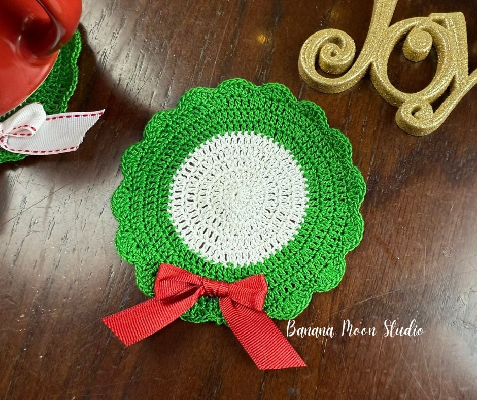Wreath shaped Christmas crochet coaster lying flat on a wooden surface with other christmas decor. Text on the image reads: banana moon studio.