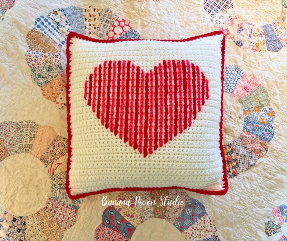 A decorative crochet pillow that is white with a red and pink heart on it and a red border. It lays on top of a quilt. Text on the image reads: Banana Moon Studio.