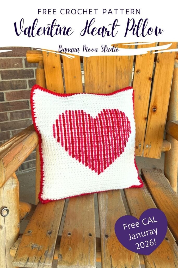 A crochet throw pillow sits on a wooden chair. The pillow is square and white with a heart crocheted on the front and a red border. Panels with text read: Free crochet pattern, valentine heart pillow, banana moon studio, free crochet along January 2026.