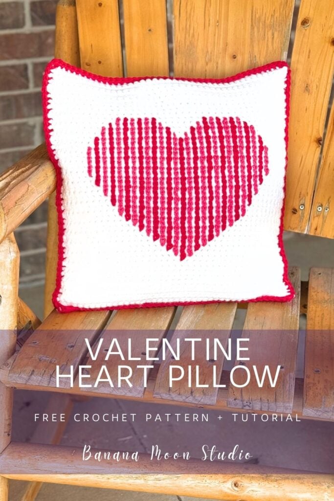 A square crochet pillow sits on a wooden chair. The pillow is white with a red and pink heart crocheted on the front and a red border. Panels with text read: valentine heart pillow, free crochet pattern + tutorial, banana moon studio.