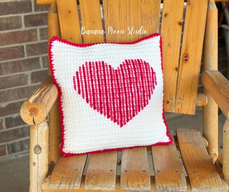 A crochet pillow sits on a wooden Adirondack chair. It is white with a heart worked across the surface. Text on the image reads: Banana Moon Studio.