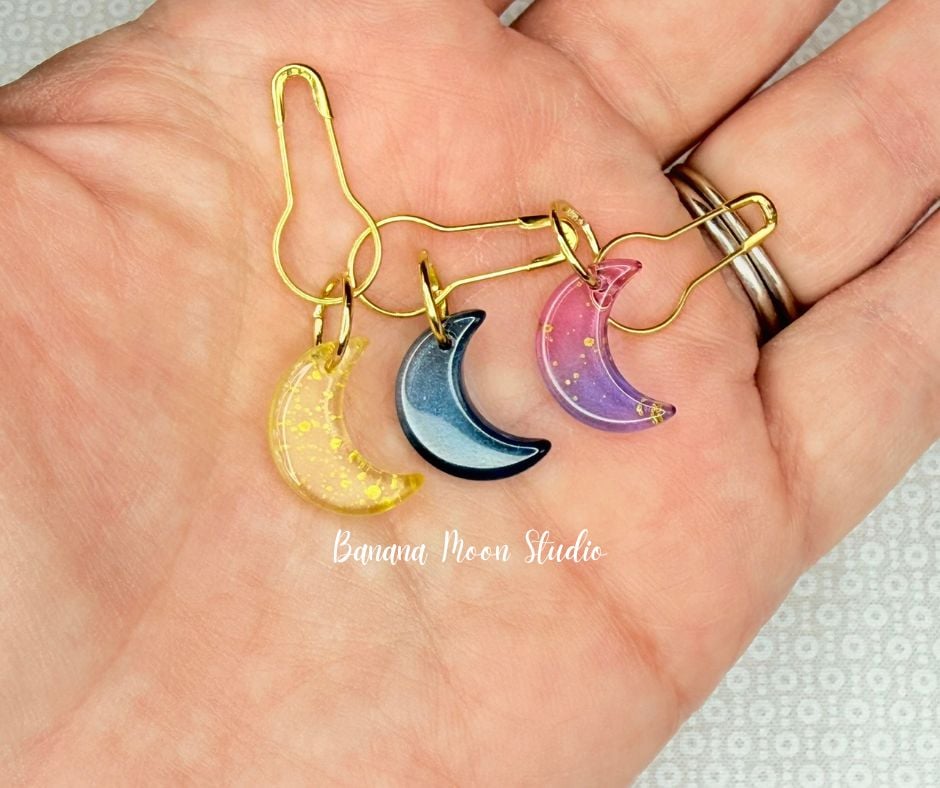 Close up of a hand holding three small stitch markers that are crescent moon charms in yellow, blue, and purple with gold colored bulb safety pins attached. Text on the photo reads: Banana Moon Studio.