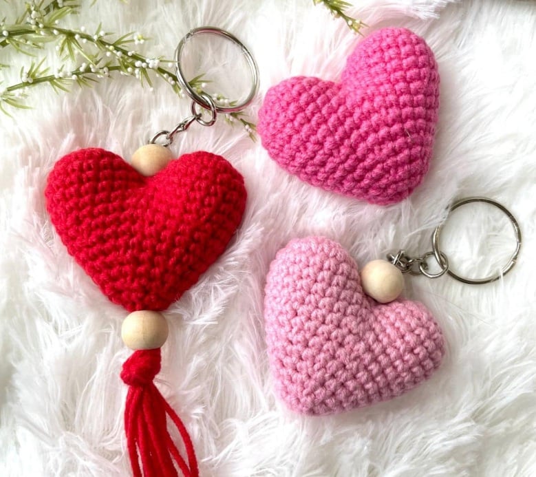 Three stuffed three dimensional hearts in pink and red on a white furry background with greenery. Two are made into keychains. 