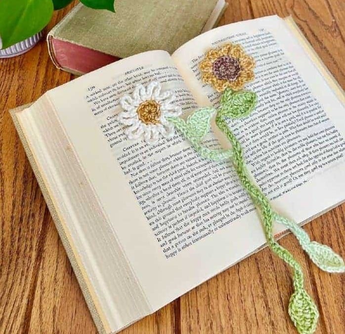 Crochet flowers with long, soft stems and leaves lie on top of an open book and can be used as bookmarks.