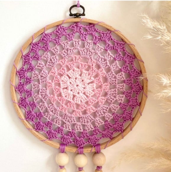 Granny stitch crochet dreamcatcher in a wooden hoop in pinks and purples hangs on a wall.
