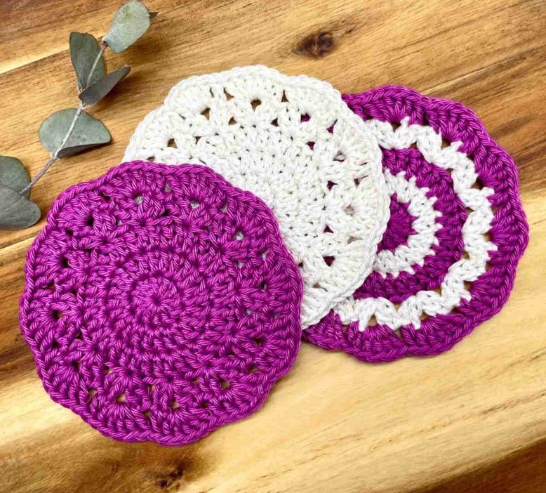 Crochet coasters that look like flowers in purple and white on a wooden surface. 
