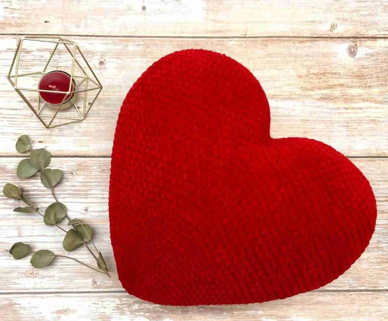 A crochet heart cushion in red chenille yarn on a wooden surface with greenery and a candle.