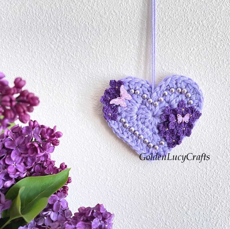 A purple crochet heart with beads, flowers, and tiny butterflies hanging against a white wall next to flowers. Text on the image reads: Golden Lucy Crafts.