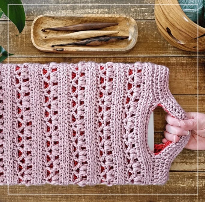 A hand holding a crochet laptop sleeve by the handles at one end and a laptop inside. It's on a table with a dish of crochet hooks.