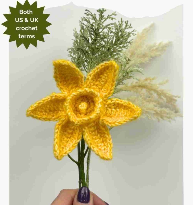 Crochet daffodil flower on a stem with greenery held up by a hand. Words on the photo read: Both US and UK crochet terms.
