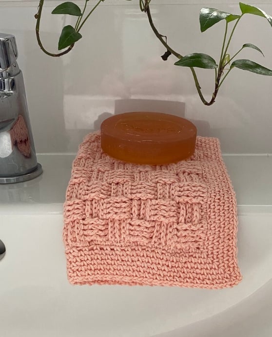 A basketweave stitch washcloth in peach folded over the edge of a bathtub with a bar of soap on top.