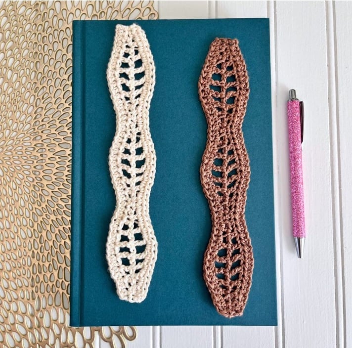 Two wavy, lacy crochet bookmarks lie on top of a teal book next to a pen.
