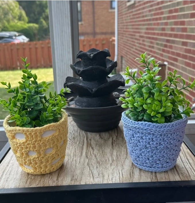 Two pots on a table with a small fountain. Each pot is covered in a crochet cozy.