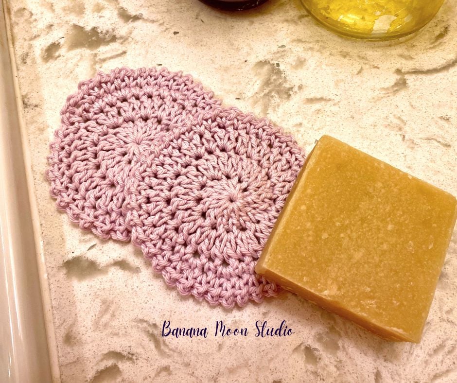 Two pale pink crochet rounds for skincare lay on a marble countertop with a bar of soap. Text on the image reads: Banana Moon Studio.