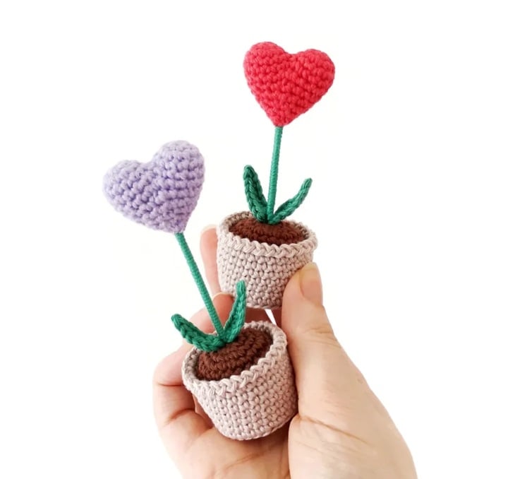 A hand holding up two little crochet hearts to look like plants sticking out of tiny crochet pots with dirt.
