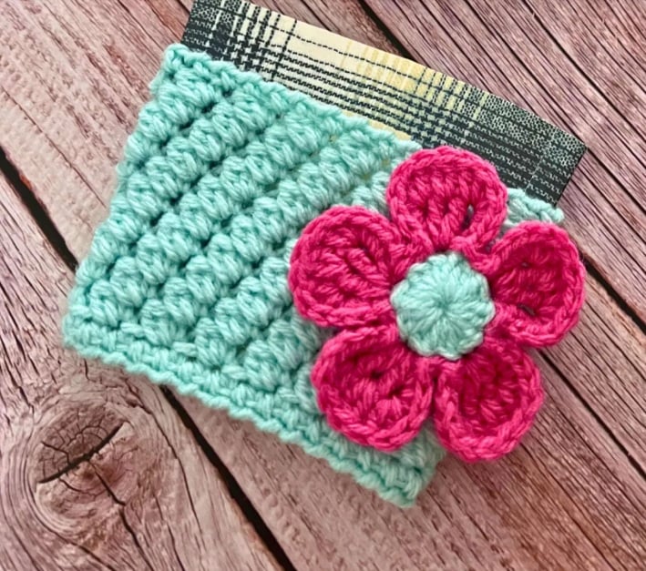 A crochet coffee sleeve in aqua with a bright pink flower on it. It sits on a wooden surface.