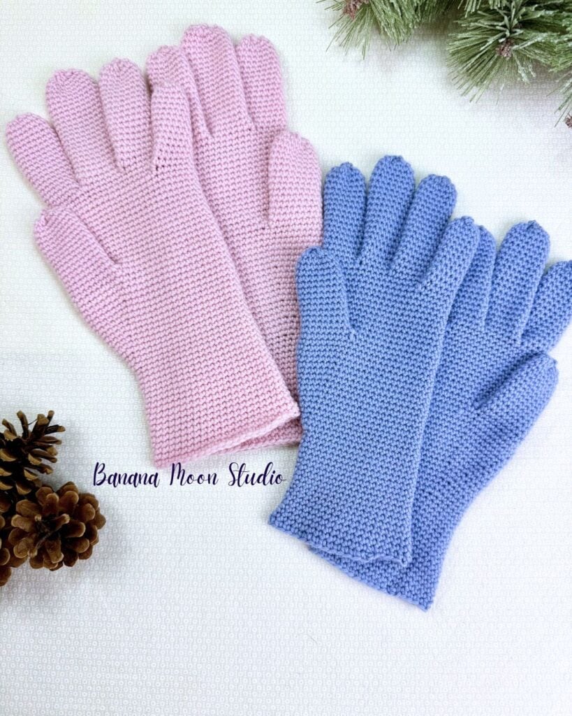 Two pairs of crochet gloves with fingers lie flat on a white surface with pinecones and an evergreen branch. Text on the image reads: Banana Moon Studio.