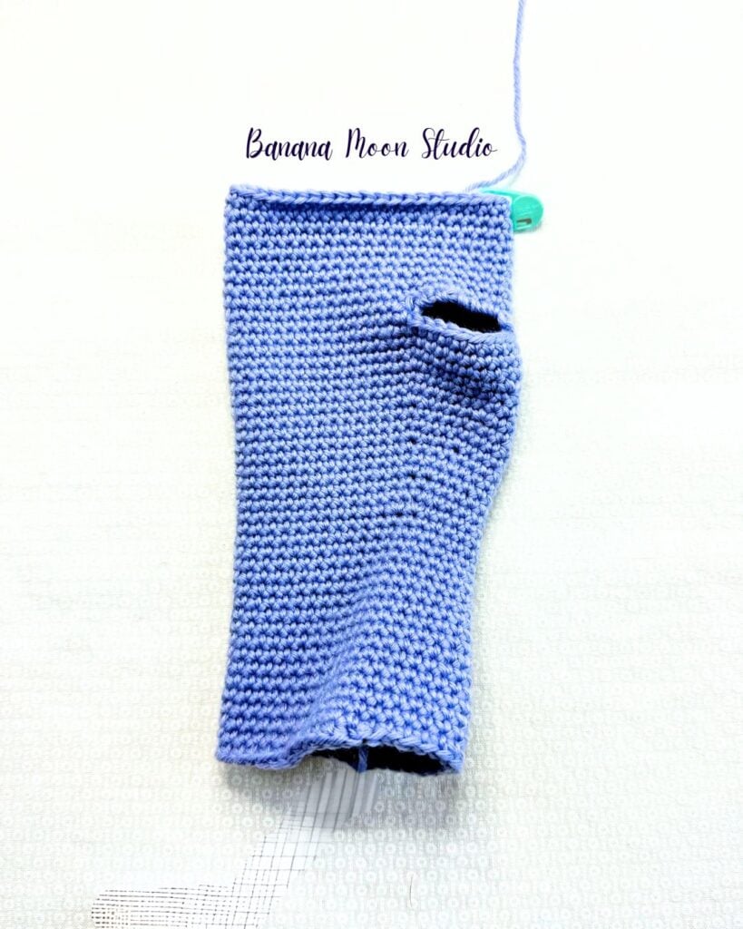 A partially finished crochet glove folded in half and lying flat on a white surface. It has a thumbhole on the right side of the glove. Text on the image reads: Banana Moon Studio.