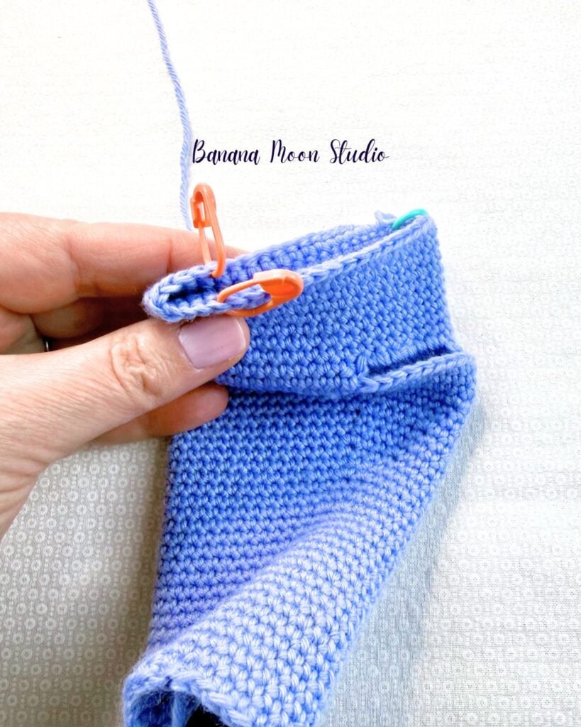 A partially finished crochet glove held by a hand and showing how to place stitch markers to mark the locations of the first and last stitch of the pinky. Text on the image reads: Banana Moon Studio.
