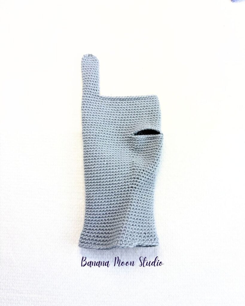 A partially finished crochet glove folded and lying flat on a white surface. This glove has a thumbhole and a pinky finger. The other fingers are missing. Text on the image reads: Banana Moon Studio.