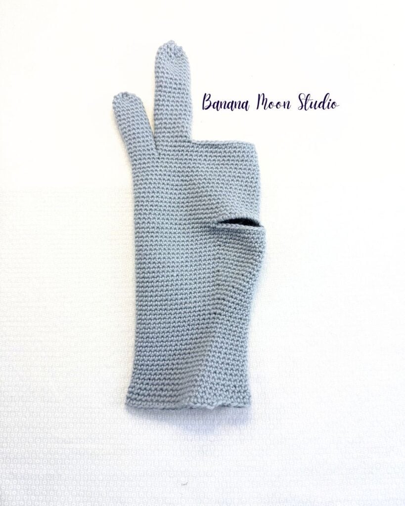 A partially finished crochet glove folded in half and lying flat on a white surface. This glove has a thumbhole, pinky finger, and ring finger. Text on the image reads: Banana Moon Studio.