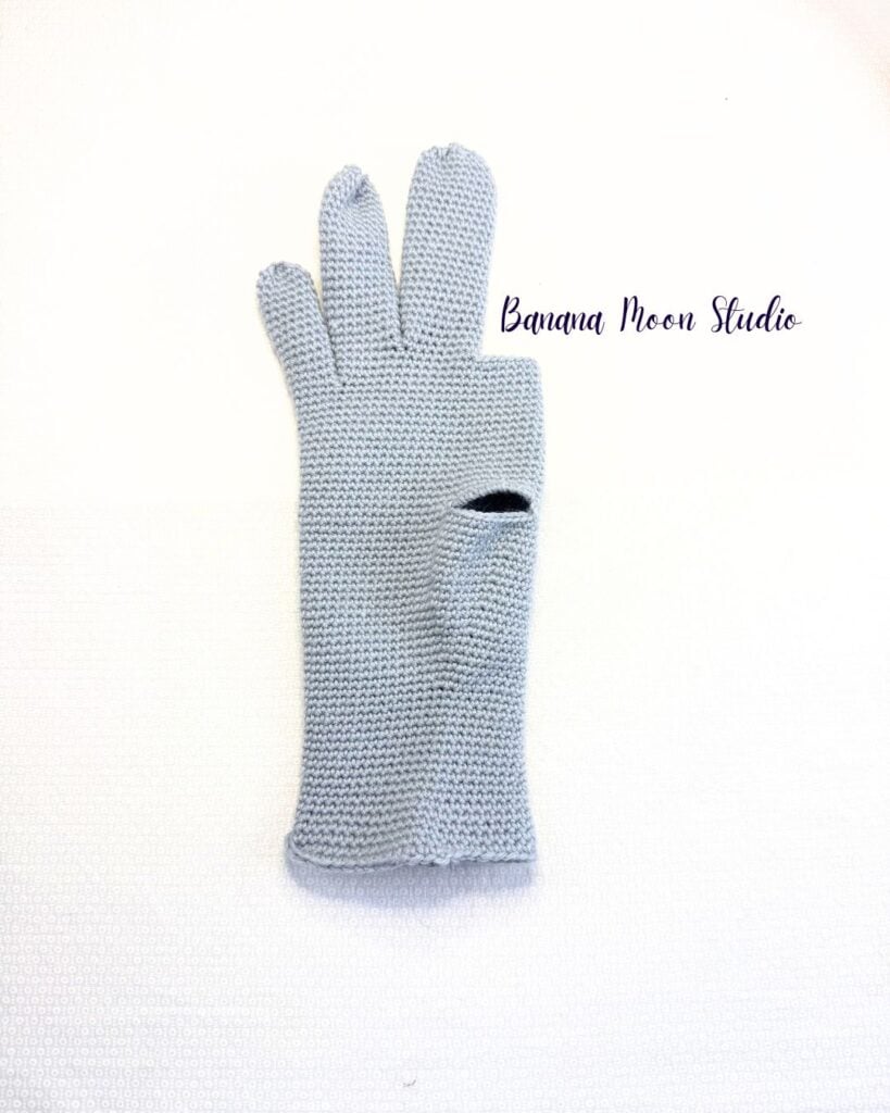 A partially finished crochet glove folded in half and lying flat on a white surface. This glove has a thumbhole, pinky finger, ring finger, and middle finger. Text on the image reads: Banana Moon Studio.