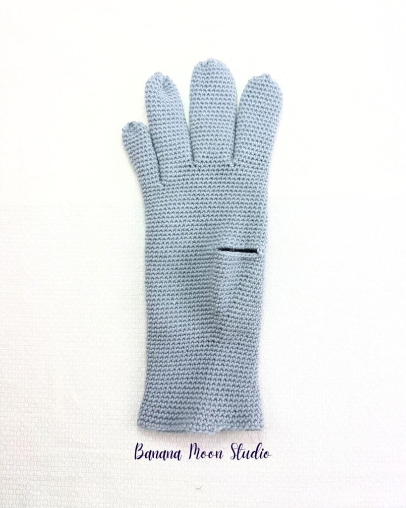 Gray crochet glove partially finished lying flat on a white surface. The glove has four fingers, but no thumb, just a thumbhole. Text on the image reads: Banana Moon Studio.