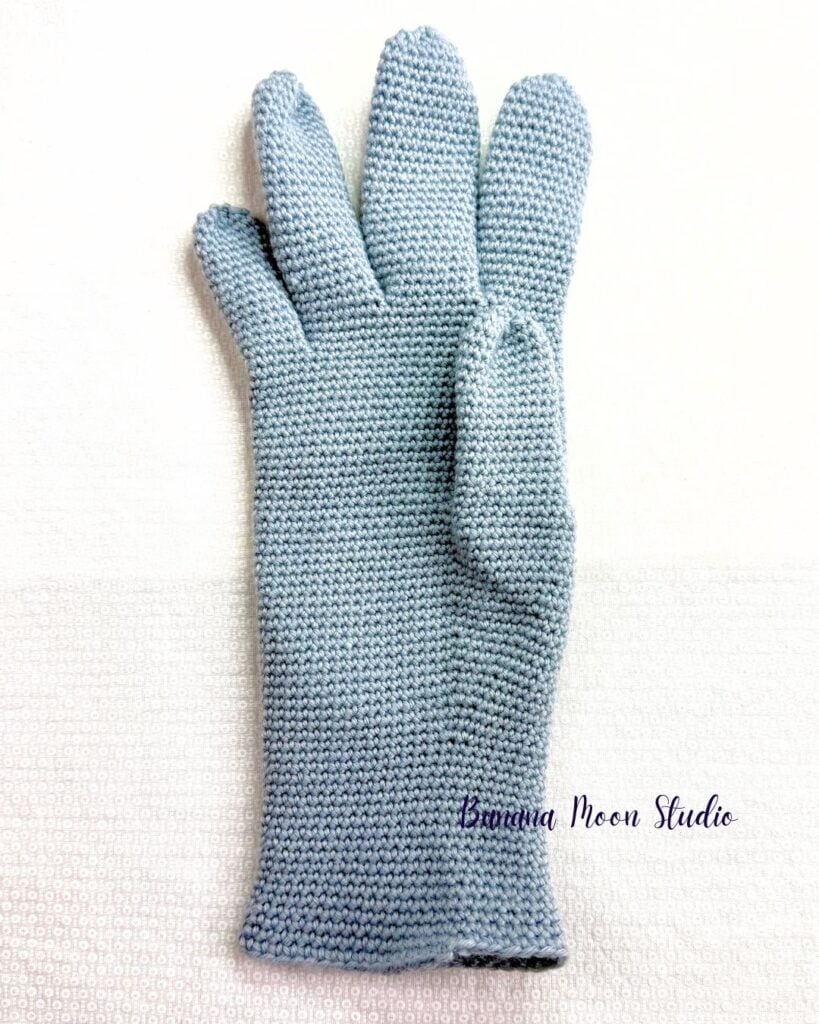 Finished gray crochet glove lying flat on a white surface. Text on the image reads: Banana Moon Studio.