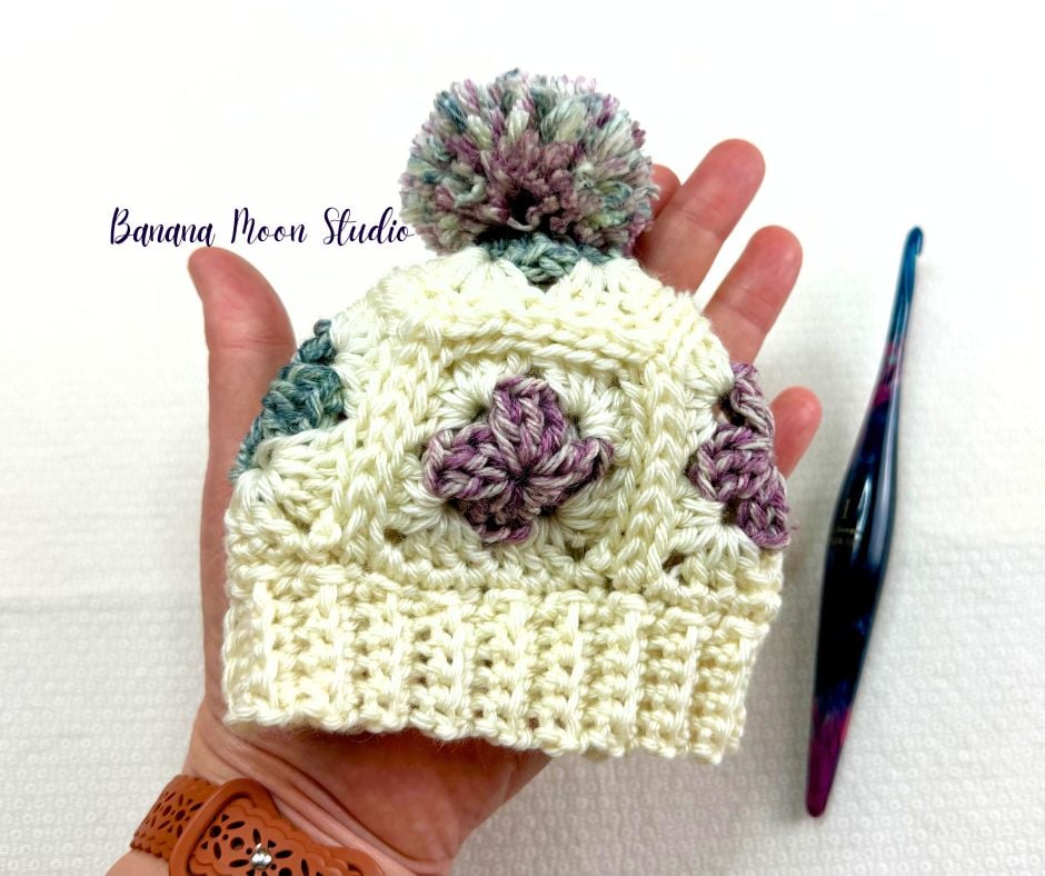 A preemie size granny square crochet hat resting on a person's hand with a crochet hook next to it. Text on the image reads: Banana Moon Studio.