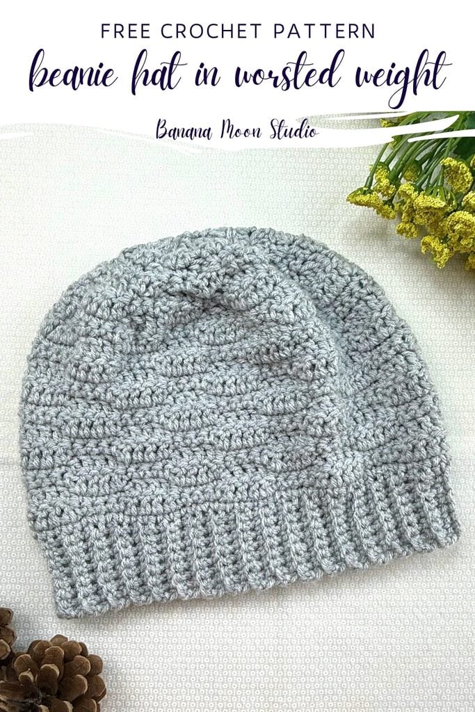 A light gray beanie lies flat on a white surface with yellow flowers. The crochet beanie has a subtle texture to it. A panel with text reads: free crochet pattern, beanie hat in worsted weight, banana moon studio.