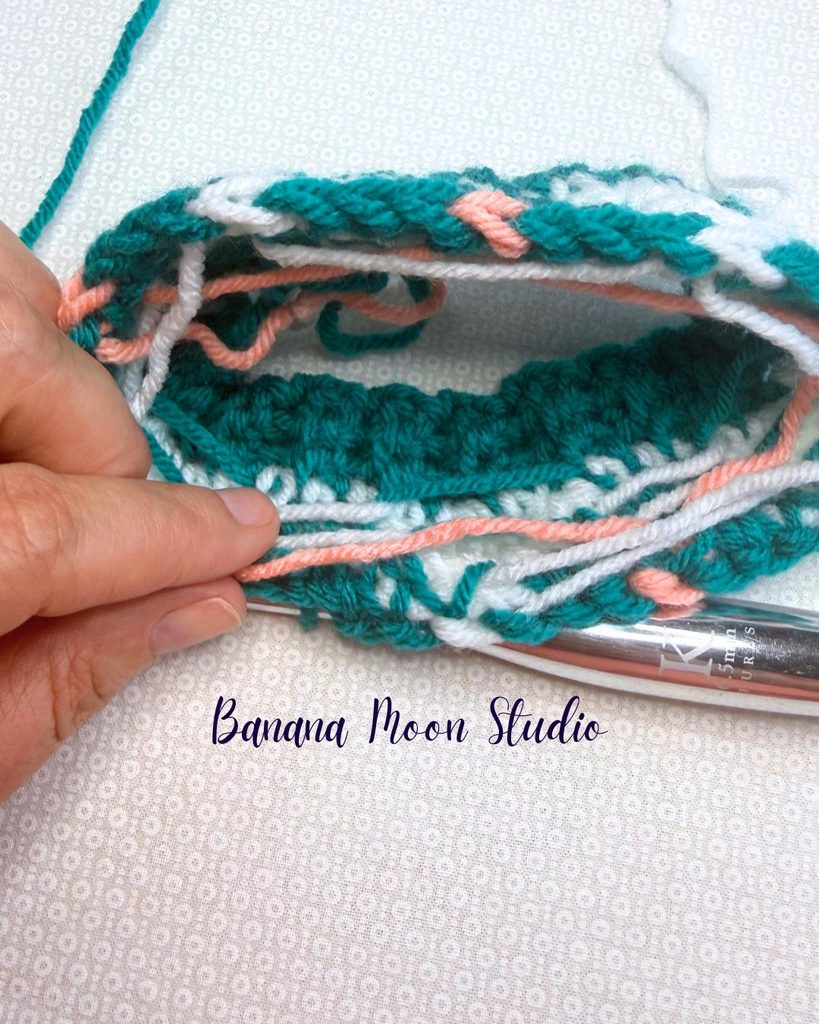Photo of a fair isle crochet hat project in process. This photo shows how to make floats across the wrong side has you work. Text on the image reads: Banana Moon Studio.