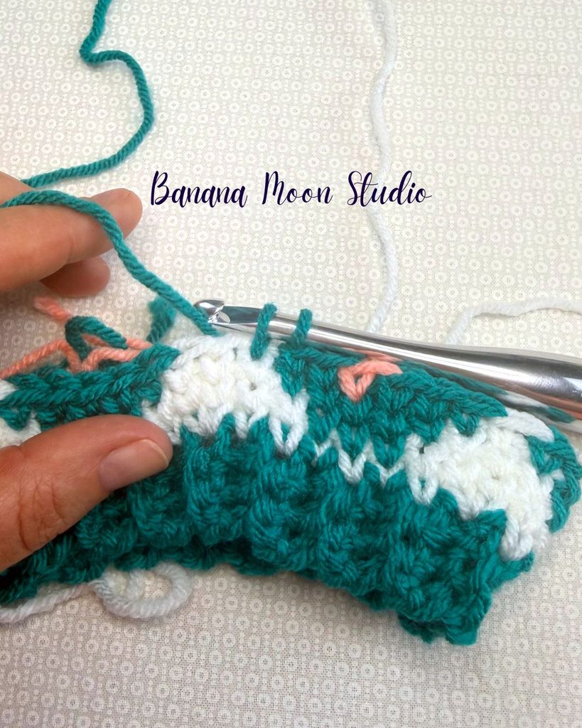 A fair isle crochet hat project in process showing a partially completed stitch with two loops on the hook. Text on the image reads: Banana Moon Studio.