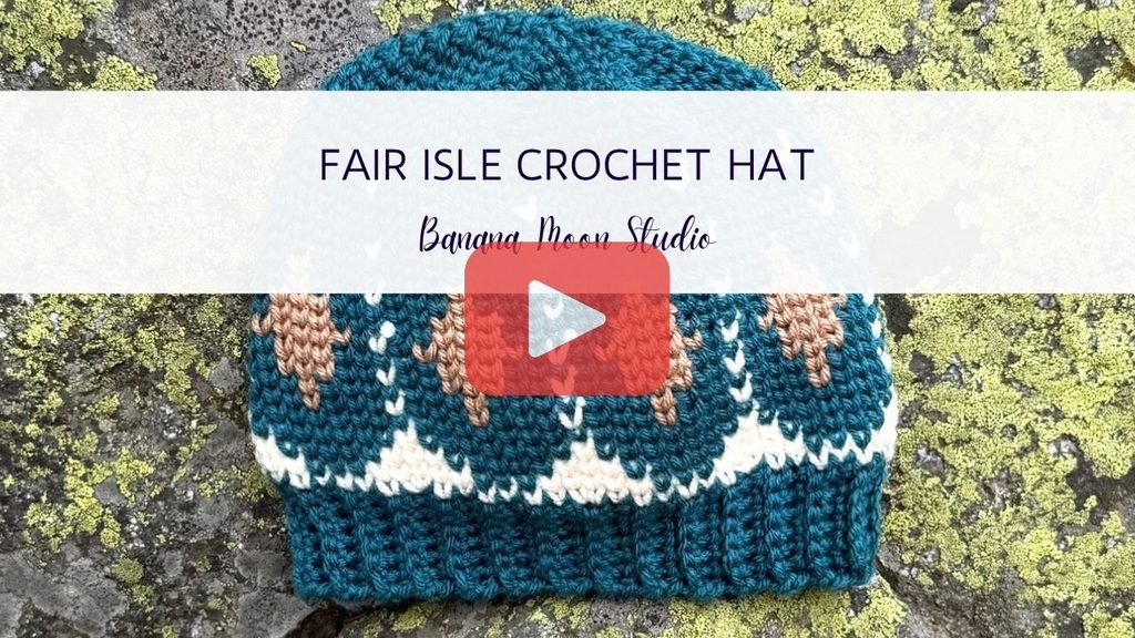 Crochet fair isle hat laying flat on a lichen covered rock. A banner with text reads: Fair Isle crochet hat, banana moon studio. A red and white play button is centered over the image.